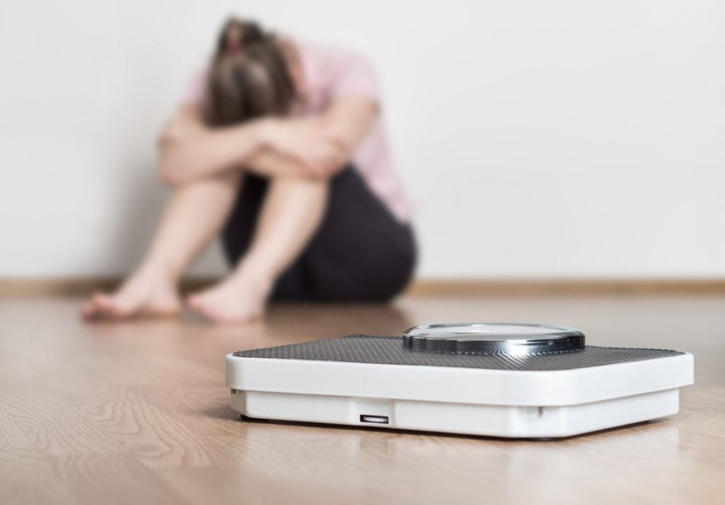 A woman with anorexia sits in a depressed pose next to a scale.