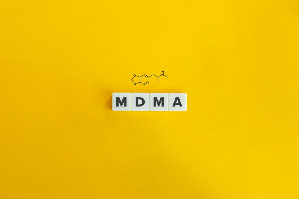 Individual scrabble tiles spelling out MDMA with a graphic of the chemical structure above it, all on a yellow background.