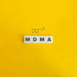 Individual scrabble tiles spelling out MDMA with a graphic of the chemical structure above it, all on a yellow background.