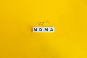 Individual scrabble tiles spelling out MDMA with a graphic of the chemical structure above it, all on a yellow background.